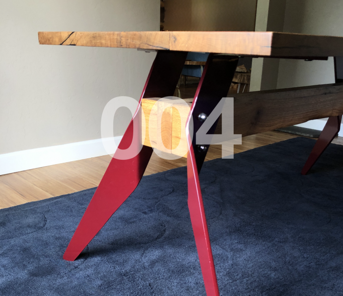 004 Table. Perspective image of a dining table. Top of table is rectangular oak. The legs are maroonish-red powder coated aluminum and bent out in a triangular fashion. There is a center plank of oak that connects the legs on each side of the table.