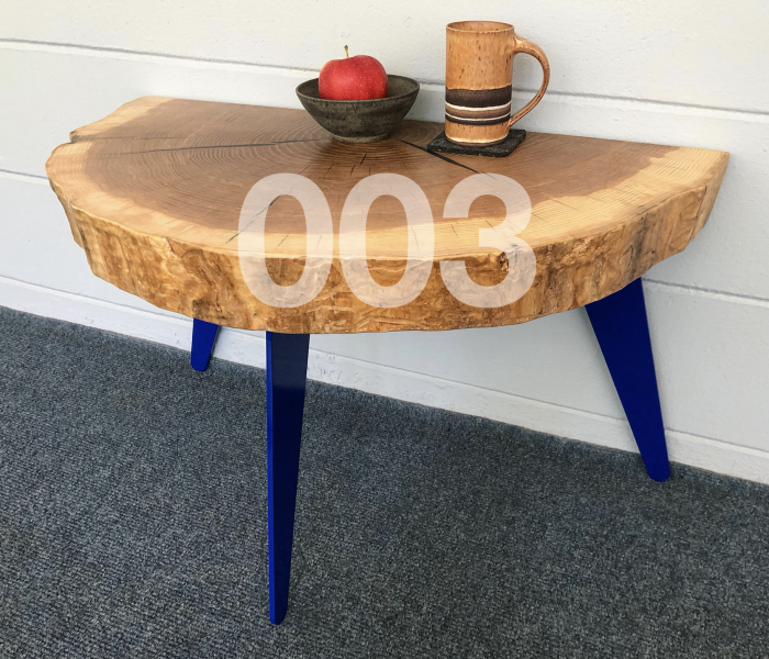 003 table. coffee table with a live edge thick slab of ash wood with light stain. The table has three legs that angle out slightly and are blue in color. There is a brown striped coffee cup and a dark grey bowl with a red apple in it on the table.