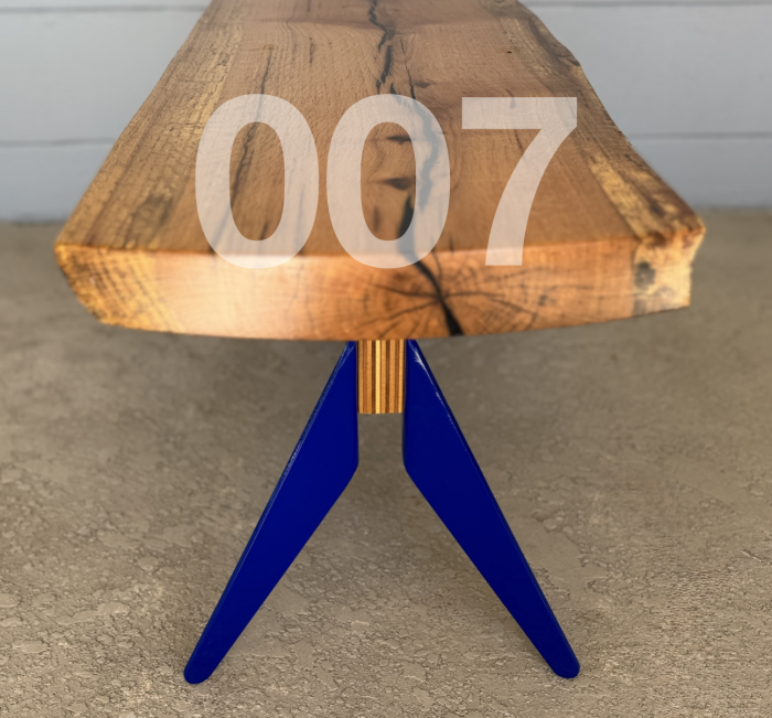 007. Image of a bench. Top of bench is a live edge piece of oak. The legs are blue powder coated aluminum. and bent out in a triangular fashion. There is a center piece of laminated plywood that connects the legs on each side of the bench.