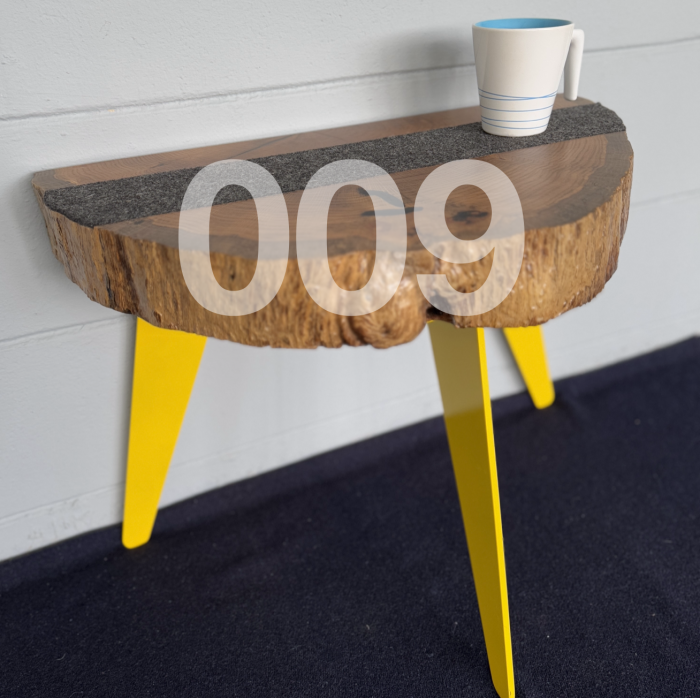 009 table. Coffee table with a thick slab of live edge oak wood with light stain. The table top is roughly a half circle shape with a thin strip of dark grey wool running parallel to the straight edge of the top. The table has three legs that angle out slightly and are yellow in color. There is a white coffee cup with thin blue stripes on the cup on the table.