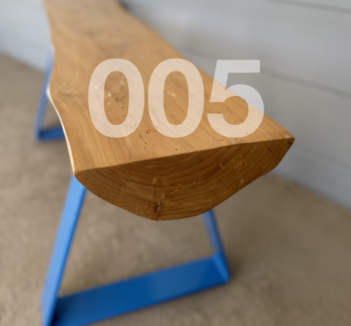 005 bench. Angled close up view of a bench. The top is a section of an ash log - flat on top and curved on the bottom the bench top. The top is held up by two bent loops of steel bar that has been powder coated light blue.