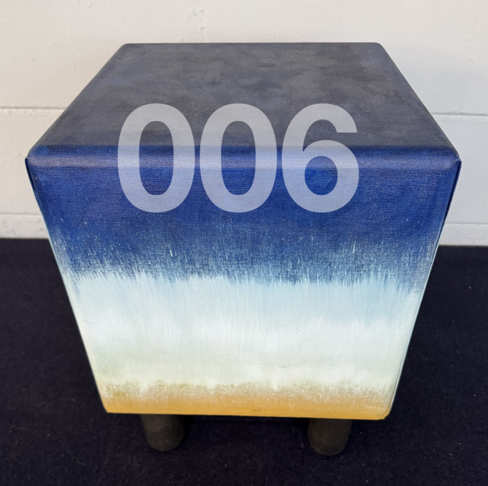 Image of a cubic stool with number 006 in front. stool has short black lesson the bottom. Entire stool is wrapped in canvas and painted. The painting is blue on top, white in the middle, and goldenrod color on the bottom.